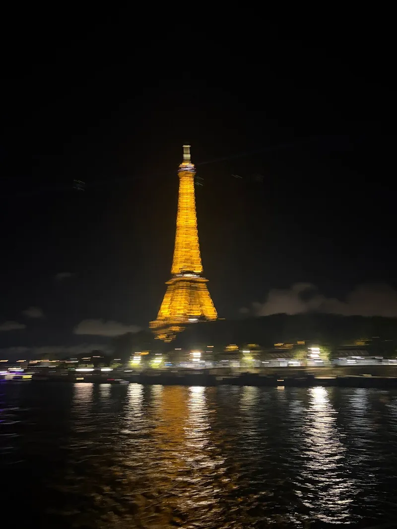 Ein unscharfes Foto des golden beleuchteten Eiffelturms und seiner Spiegelung, nachts vom Fluss aus aufgenommen